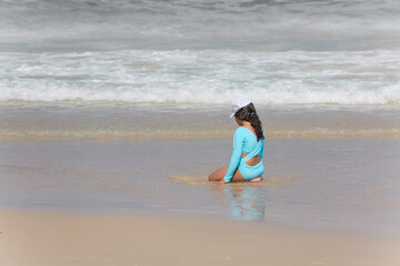 A teenage girl in a blue swimsuit is sitting on the seashore