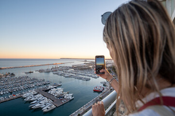 Fototapeta na wymiar Woman takes a photo of the Port of Alicante from a hotel terrace in Spain.