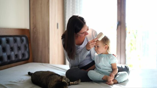 Mom combs a little girl sitting on a bed near a lying cat