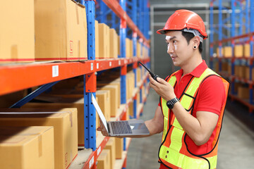 Warehouse workers man with hardhats and reflective jackets using computer, walkie talkie radio and cardboard while controlling stock and inventory in retail warehouse logistics, distribution center