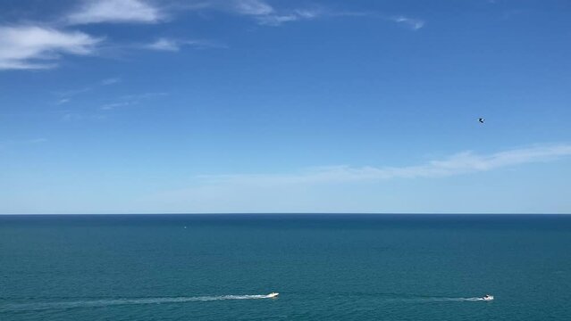 Pristine view from Piazza Nuova of Numana at the Adriatic Sea with its deep blue rippling waters, two white boats skimming to the right and a bird flying in the opposite direction under a dreamy sky