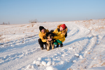 Fototapeta na wymiar Male with female in yellow coats walking with big black dog on winter background. Family winter activity with pet on sunny day outdoor. 
