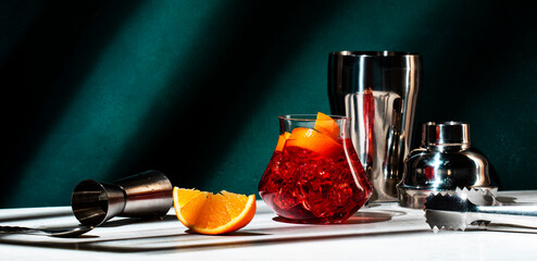 Boulevardier alcoholic cocktail with bourbon, vermouth, bitters, orange and ice. Dark green background, hard light and shadow pattern. Banner