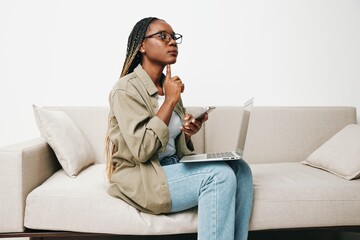 Fototapeta na wymiar African American woman business freelancer working sitting on the couch at home in a laptop and phone, business calls and correspondence sadness and anger, home clothes and eyeglasses, light interior