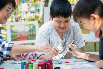 Learning to use watercolor to draw various pictures of a young Southeast Asian boy on a table outside classroom with his female art teacher, raising teen and adult helps kids school project concept.