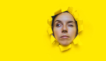Close-up portrait of a caucasian young woman looking through a hole in yellow paper and winking. An incredulous look. Women's curiosity and gossip. Jealous wife. Spy concept.