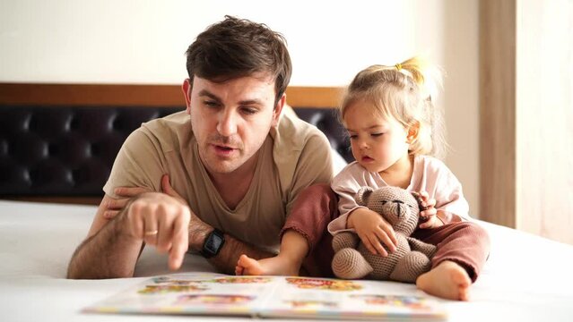 Father reading picture book to little girl while lying on bed