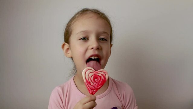 A happy girl happily licks a heart-shaped lollipop. delicious dessert. Valentine's Day. Isolated white background