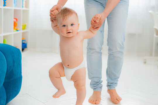 Cute little baby in a diaper learning to walk in a white room , mom holding his hands