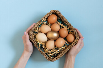 Gift basket with easter eggs. Women hands holding golden and natural color eggsin basket. Top view on pastel blue background.