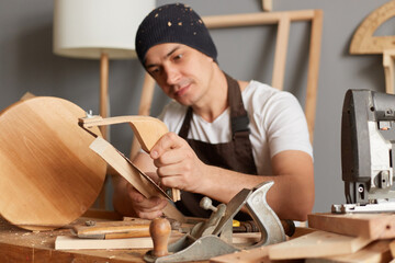 Image of calm attractive young adult man carpenter wearing white t-shirt, black cap and brown apron working in joinery, making wooden chair, sanding wood block.