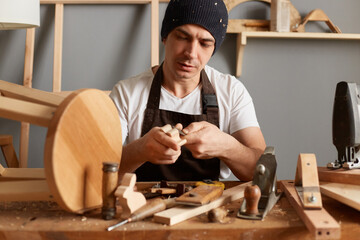 Portrait of concentrated young adult man carpenter wearing white t-shirt, black cap and brown apron working, joinery work, wood carving, chisels for carving on the woodworker desk timber joinery work.