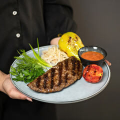 Waiter in black uniform serves lunch. Grilled meat steak with vegetables, herbs and sauce on plate in hands of woman. Side view. Close-up. Soft focus. 