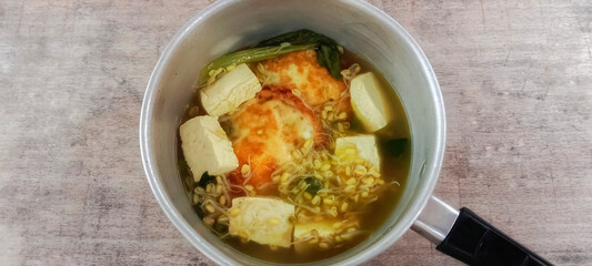 Homemade tofu soup, eggs, bean sprouts and mustard greens served in a pot against a rough textured wooden background