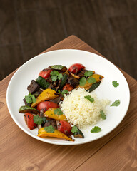 Baked meat with vegetables and boiled rice for garnish. Dish is decorated with parsley in white plate on wooden table. Complex lunch. View from above. Copy space. Soft focus. 