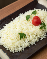 Bowl of boiled long-grain basmati rice. Rectangular black plate with dish on wooden table. Rice is garnished with herbs and cherry tomatoes. Wooden table. View from above. Copy space. Close-up.