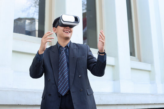 Low angle shot of attractive young businessman wearing suit and VR glasses outside the office, smiling during online simulation and gesture touching.  - Powered by Adobe