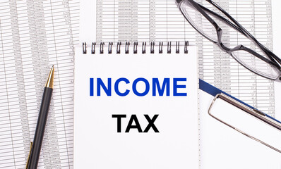 A white notepad with the text INCOME TAX a pen, reports and glasses lie on a white office table....