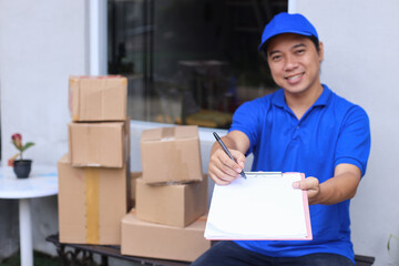 Young deliveryman showing shipping document to be signed while sitting with pile of boxes to send to customers outside the office.