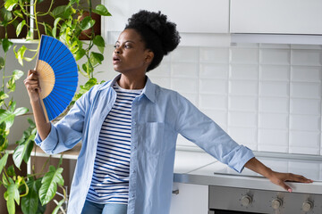 Overheating concept. African American woman fanning herself with hand fan experiencing health problems due to hot weather. Ethnic black girl looks into distance standing in room with home plants