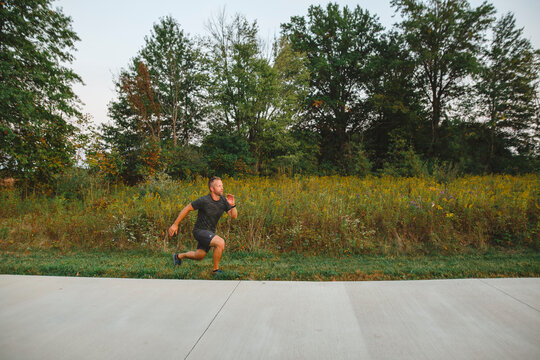 A fit male warms up with lunges along tree-lined path in park at dusk