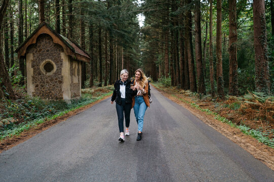 Two women walking on a road in the forest while watching their phone