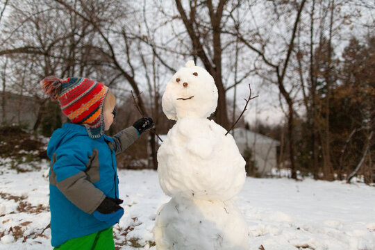 Toddler boy in cold weather gear putting stick arm into a snowman