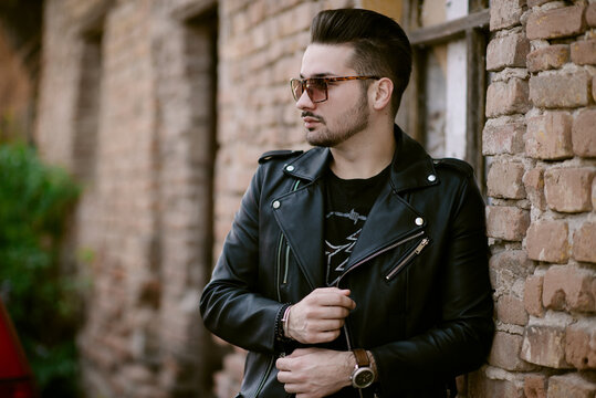 Trendy Man with sunglasses in a jacket standing by a brick wall.