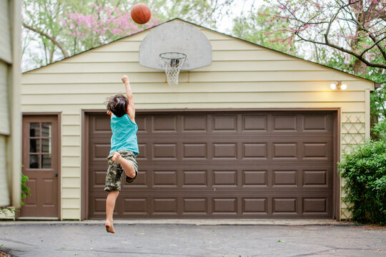 A barefoot boy leaps up to shoot a basket in a driveway at dusk