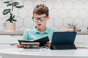 Portrait of young preteen focused boy wearing blue T-shirt, glasses, sitting at desk near notepads,...