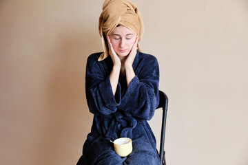 woman in a robe and with a towel on her head applies cosmetics to skin