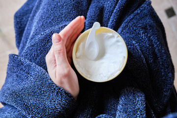 woman in a robe and with a towel on her head applies cosmetics to skin