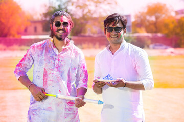 Portrait of happy young Indian men wearing white kurta holding plate full of powder color and pichkari celebrating holi festival at park outdoor, Face painted with colorful gulal