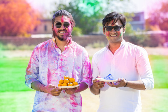 Portrait of happy young Indian men wearing white kurta holding plate full of powder color celebrating holi festival at park outdoor, Face painted with colorful gulal