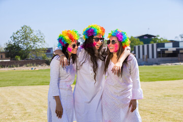 Group of playful young indian women wearing white kurta and colorful hair wigs celebrating holi...