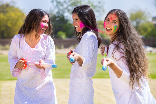 Group of young indian girl friends wearing white kurta outfit playing with pichkari celebrating holi festival outdoor at park.