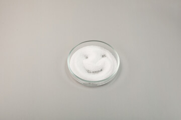 Sugar substitute in petri dish on grey background. White crystalline powder in the shape of smiley...