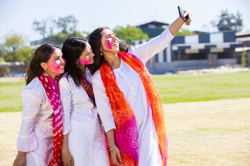 Portrait of happy young indian female friends taking selfie picture with smart phone while celebrating holif festival together at park.