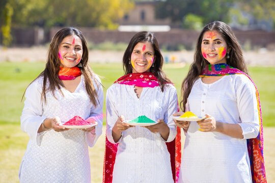 Group of happy young Indian women wearing white cloths holding powder color plate celebrating holi festival at park outdoor, Face painted with colorful gulal. Looking at camera.
