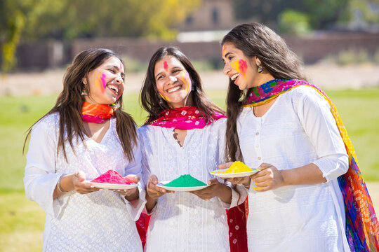 Group of young happy indian woman wearing white kurta dress holding color powder plates in hand celebrating holi festival together at park with their face painted colorful gulal