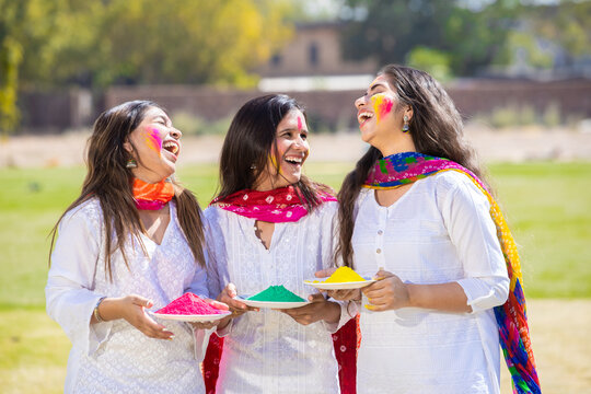 Group of happy indian friends wearing white kurta dress holding color powder plates in hand celebrating holi festival together at park with their face painted colorful gulal