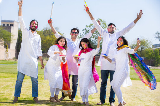 Group of cheerful young Indian family celebrating holi festival at park outdoor, Playful adult male and friends with colorful paint faces playing with gulal.