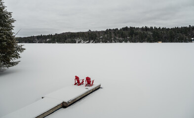 Red adirondack chairs on end of snowy dock on a lake in winter.