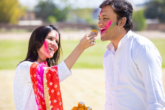 Happy young indian couple celebrating holi festival together while enjoying delicious laddu sweet.