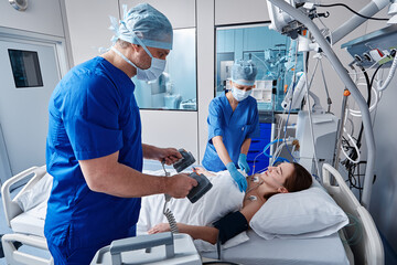 Doctor holding defibrillator on unconscious female patient's chest in intensive care unit during...