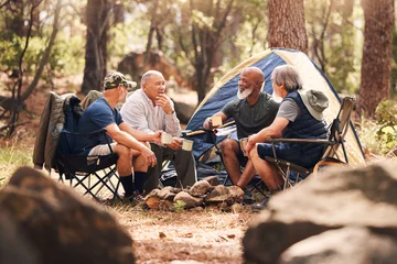 Foto op Plexiglas Senior people, camping and relaxing in nature for travel, adventure or summer vacation together on chairs by tent in forest. Group of elderly men talking, enjoying camp out conversation in the woods © Azee J/peopleimages.com