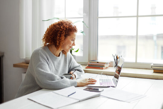 Side view of pretty young redhead teacher sitting at table next to huge window at university, drinking coffee and checking e-mail during brake, having rest between lessons, preparing material