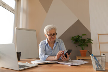 Fototapeta na wymiar Confident stylish european middle aged senior woman using smartphone at workplace. Stylish older mature 60s gray haired lady businesswoman with cell phone in office. Boss leader using internet apps