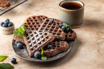 Heart form chocolate waffle cookies in ceramic plate decorated with berries, mint and powdered sugar with tea for Valentine's day on beige background. Sweet gift with love, text space