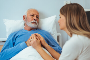 Fototapeta premium In the Hospital Ward Recovering Father is Visited by Daughter. Senior Sick Man Sleeping in Bed Daughter Sits Beside, Worrying. Modern Private Ward. Family Values. 